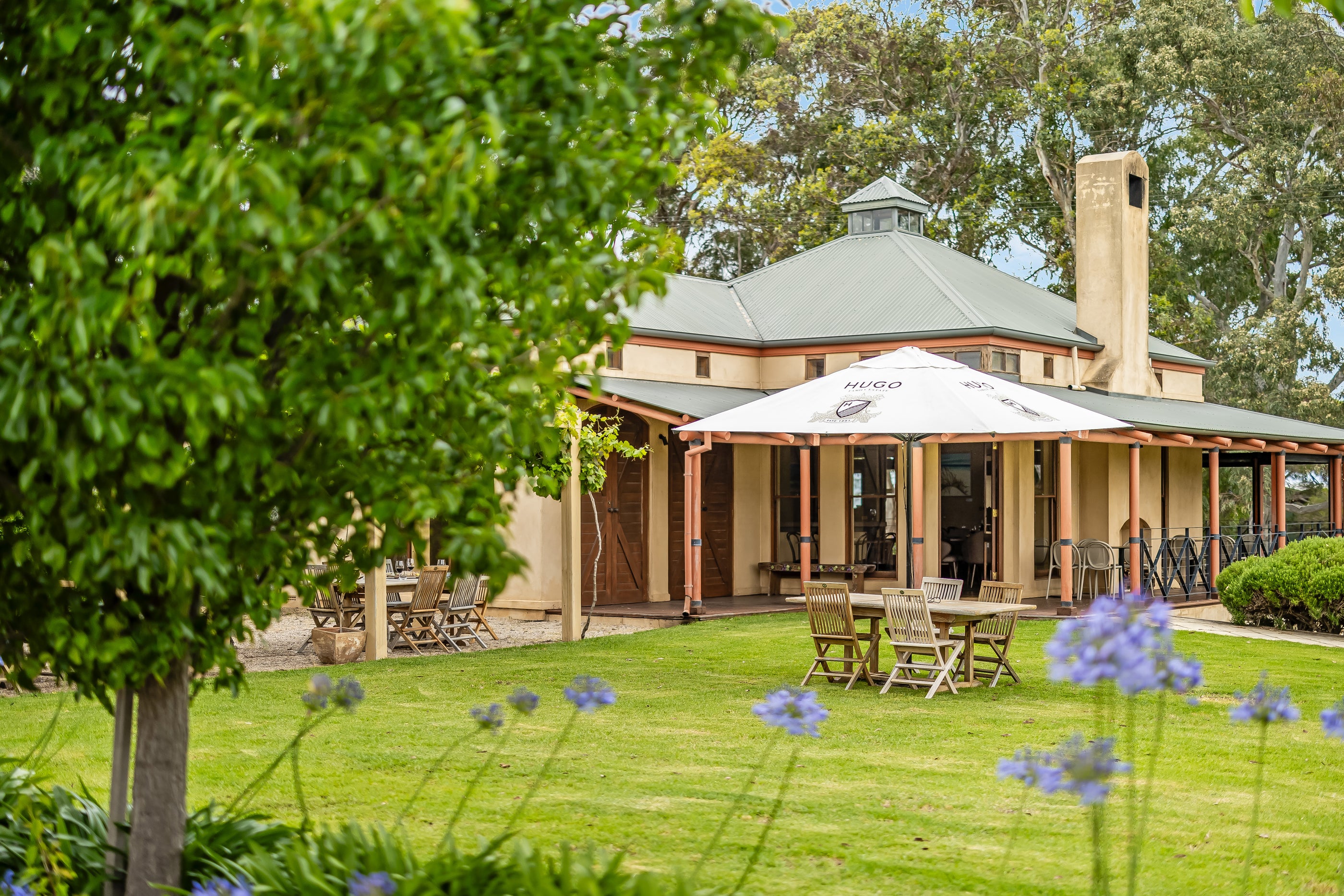 Hugo Wines, outdoor setting with a building, tables, chairs, and an umbrella on a grassy area with trees and flowers.